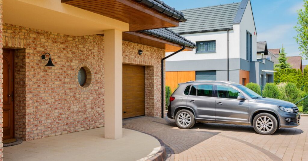 A gray SUV sits in the driveway of a brick home, parked just outside the garage on a bright, sunny day.