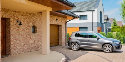A gray SUV sits in the driveway of a brick home, parked just outside the garage on a bright, sunny day.