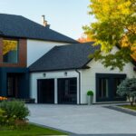 Modern white and dark gray house with a wide paved driveway, trimmed lawn, shrubs, and trees in warm light.
