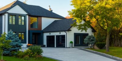 Modern white and dark gray house with a wide paved driveway, trimmed lawn, shrubs, and trees in warm light.