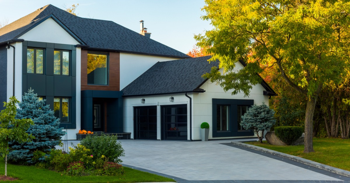 Modern white and dark gray house with a wide paved driveway, trimmed lawn, shrubs, and trees in warm light.
