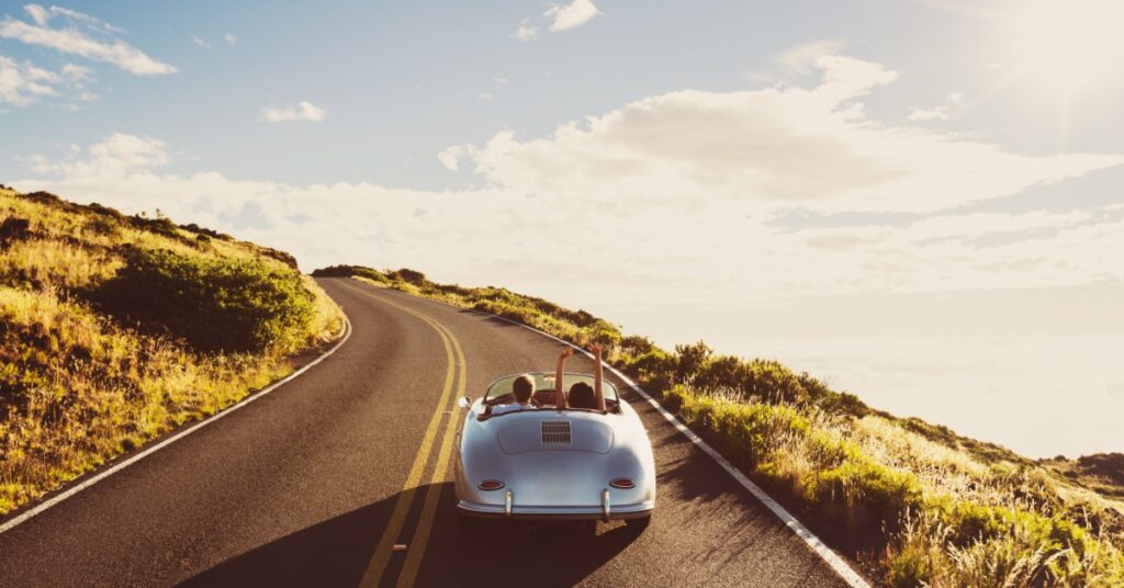 Silver convertible with two passengers drives up a winding hillside road under bright sun and scattered clouds.
