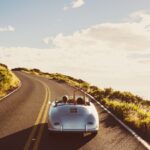 Silver convertible with two passengers drives up a winding hillside road under bright sun and scattered clouds.