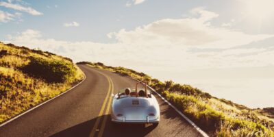 Silver convertible with two passengers drives up a winding hillside road under bright sun and scattered clouds.