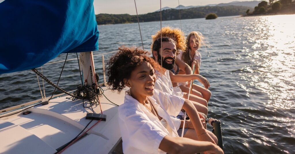 Friends laugh together on a sailboat, leaning over the side as sunlight sparkles on a lake and hills rise beyond.
