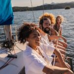 Friends laugh together on a sailboat, leaning over the side as sunlight sparkles on a lake and hills rise beyond.