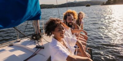 Friends laugh together on a sailboat, leaning over the side as sunlight sparkles on a lake and hills rise beyond.