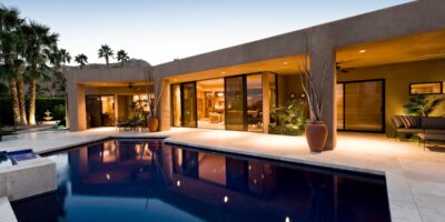 A close-up of the backyard space of a modern, minimalist-style home. There is a pool and two covered seating areas.