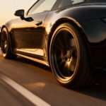 A black sports car driving down a road at sunset with the sky yellowing in the distance. The lines on the road are blurred.