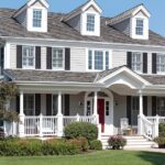 Large gray two-story house with white trim, black shutters, three dormers, a wide porch, and a manicured lawn.