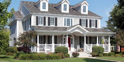 Large gray two-story house with white trim, black shutters, three dormers, a wide porch, and a manicured lawn.