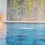 What Types of Spas Can You Choose for Your Property? A woman resting in a blue outdoor pool beside a folded towel and flower, with plants and a textured wall nearby.