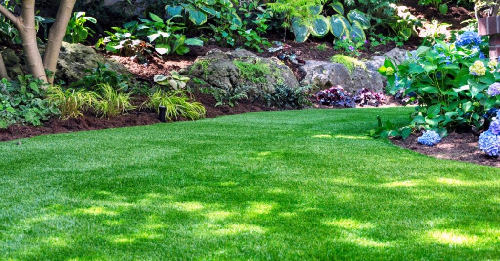 A green artificial lawn bordered by leafy garden plants, shrubs, mulch, and trees in a small backyard garden.