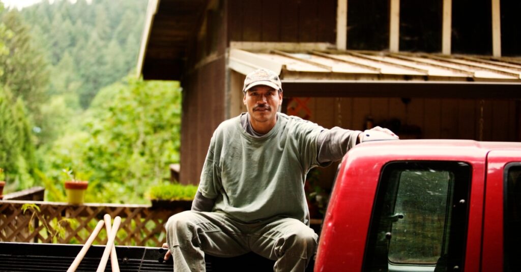 A man sitting in the bed of a red pickup truck beside a wooden building with trees and plants behind him.