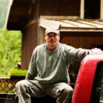Truck Upgrades That Support Work and Play A man sitting in the bed of a red pickup truck beside a wooden building with trees and plants behind him.