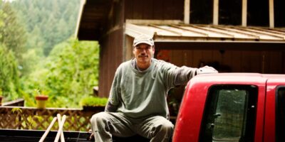 A man sitting in the bed of a red pickup truck beside a wooden building with trees and plants behind him.
