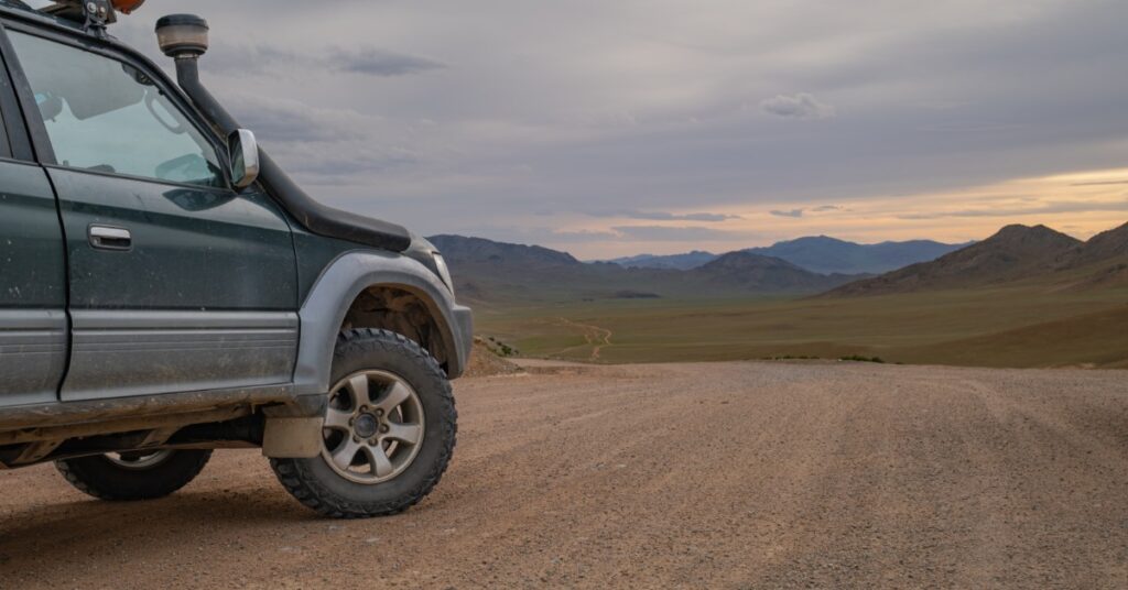 A dark SUV is parked on a dirt road in a wide desert valley, with rocky hills under a cloudy sky as the sun sets.