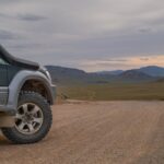 A dark SUV is parked on a dirt road in a wide desert valley, with rocky hills under a cloudy sky as the sun sets.