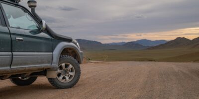 A dark SUV is parked on a dirt road in a wide desert valley, with rocky hills under a cloudy sky as the sun sets.