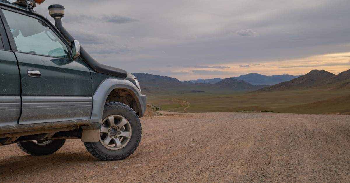 A dark SUV is parked on a dirt road in a wide desert valley, with rocky hills under a cloudy sky as the sun sets.