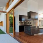 A modern luxury home features expansive glass walls, warm wood floors, and open views of a green backyard.