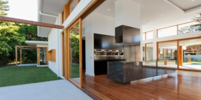 A modern luxury home features expansive glass walls, warm wood floors, and open views of a green backyard.