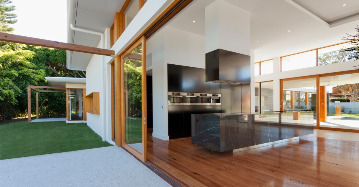 A modern luxury home features expansive glass walls, warm wood floors, and open views of a green backyard.