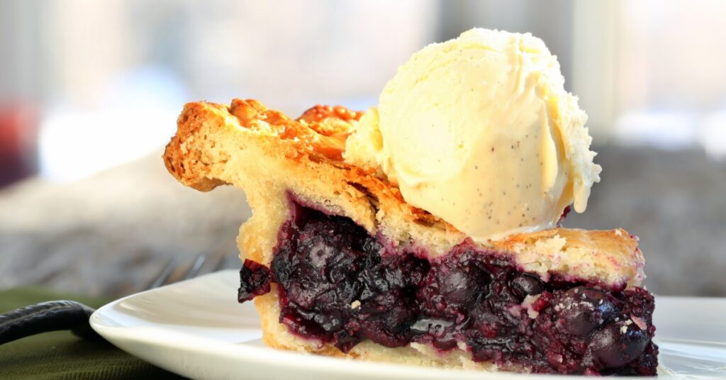 A slice of blueberry pie on a small white plate. A thick scoop of vanilla ice cream sits on top of the pie crust.