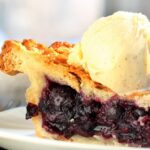 A slice of blueberry pie on a small white plate. A thick scoop of vanilla ice cream sits on top of the pie crust.