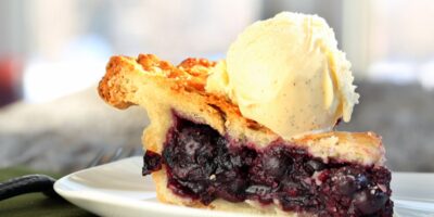 A slice of blueberry pie on a small white plate. A thick scoop of vanilla ice cream sits on top of the pie crust.