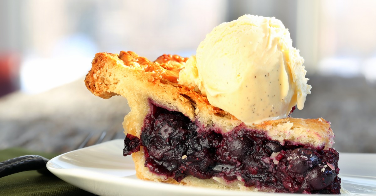 A slice of blueberry pie on a small white plate. A thick scoop of vanilla ice cream sits on top of the pie crust.