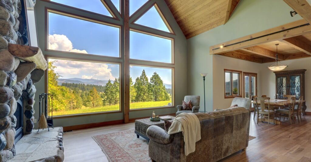 A luxury living room with large windows overlooks a wooded landscape and distant mountains beneath a partly cloudy sky.