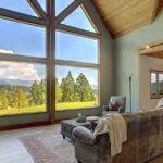 How Surroundings Shape a Luxury Home Experience A luxury living room with large windows overlooks a wooded landscape and distant mountains beneath a partly cloudy sky.