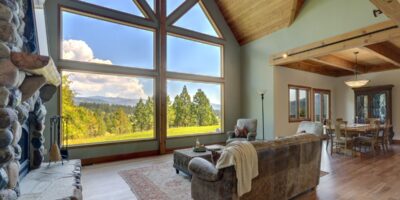 A luxury living room with large windows overlooks a wooded landscape and distant mountains beneath a partly cloudy sky.