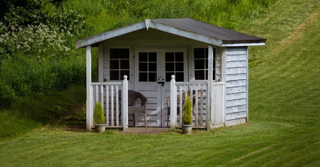 A small white garden shed with a porch, chairs, and potted plants sitting on a green lawn near shrubs.