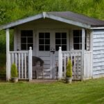 Types of Outbuildings That Can Add Property Value A small white garden shed with a porch, chairs, and potted plants sitting on a green lawn near shrubs.
