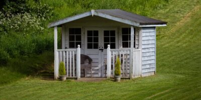 A small white garden shed with a porch, chairs, and potted plants sitting on a green lawn near shrubs.