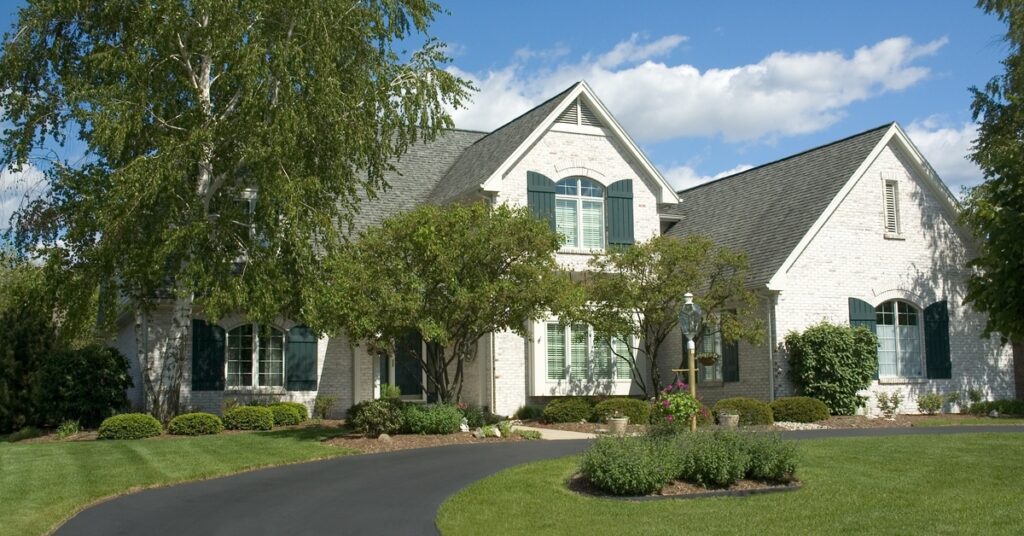 A large white brick house with green shutters, a curved driveway, trees, and a landscaped lawn under a blue sky.