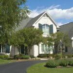 How Sealcoating Can Improve a Driveway’s Appearance A large white brick house with green shutters, a curved driveway, trees, and a landscaped lawn under a blue sky.