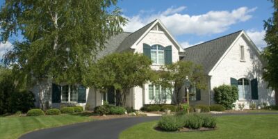 A large white brick house with green shutters, a curved driveway, trees, and a landscaped lawn under a blue sky.