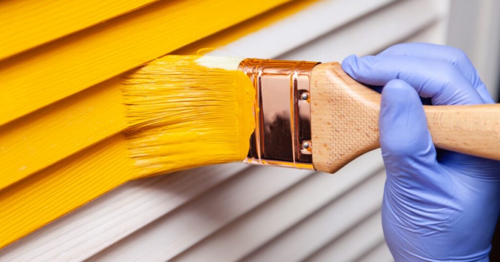A hand in a purple glove using a paintbrush to apply yellow paint onto a wooden door with visible grain.