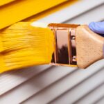 How Small Exterior Updates Can Refresh Your Home A hand in a purple glove using a paintbrush to apply yellow paint onto a wooden door with visible grain.
