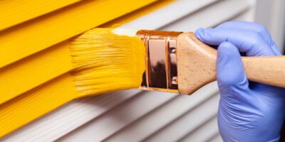 A hand in a purple glove using a paintbrush to apply yellow paint onto a wooden door with visible grain.