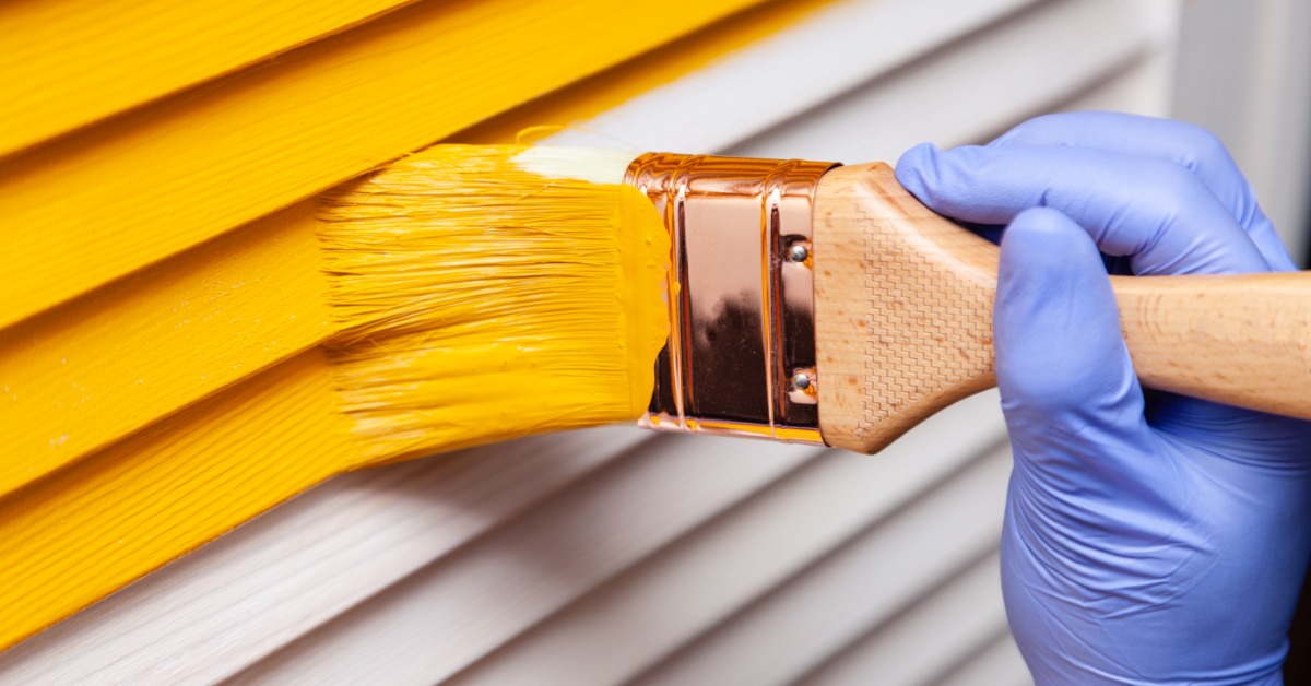 A hand in a purple glove using a paintbrush to apply yellow paint onto a wooden door with visible grain.